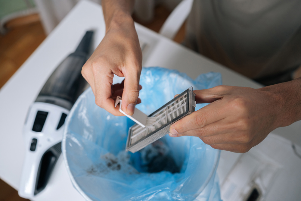 Person cleaning HEPA filter from dirty robot vacuum cleaner, carefully tapping out accumulated dust and debris into bin with blue plastic liner, performing essential device maintenance