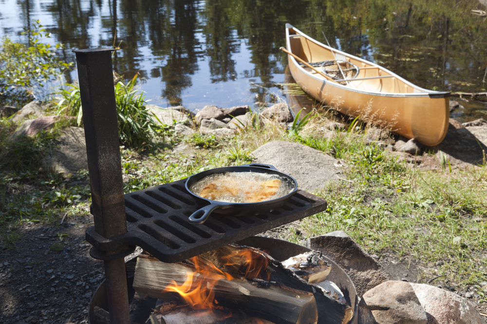 Cooking food in a cast iron skillet over an open fire with a canoe and lake in the background