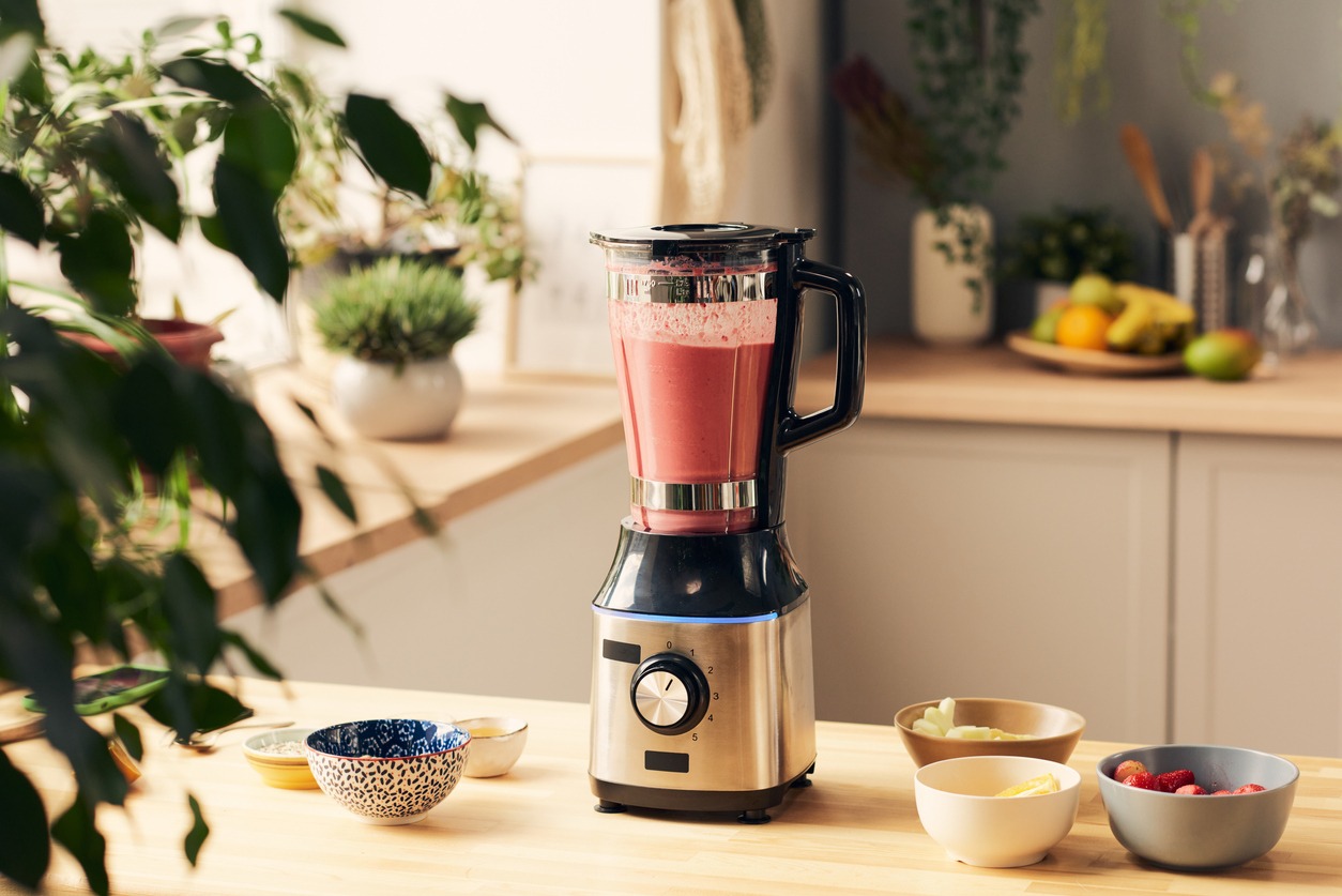 Electric blender with fresh homemade fruit smoothie and group of bowls containing ingredients on wooden kitchen table